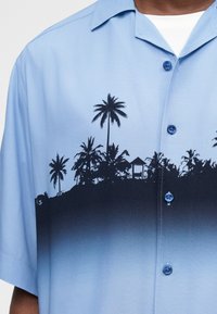 Ανοιχτό μπλε πουκάμισο με κουμπιά, με μαύρη τροπική σιλουέτα φοίνικων και μια μικρή καλύβα στο ύψος του στήθους και κοντά μανίκια.