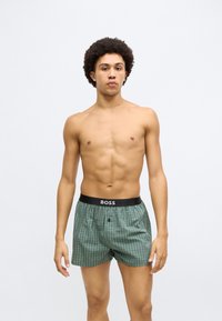 Zöld-fehér kockás boxeralsó fekete derékszalaggal, amelyen "BOSS" logó látható, egy ing nélküli férfi modell viseli.