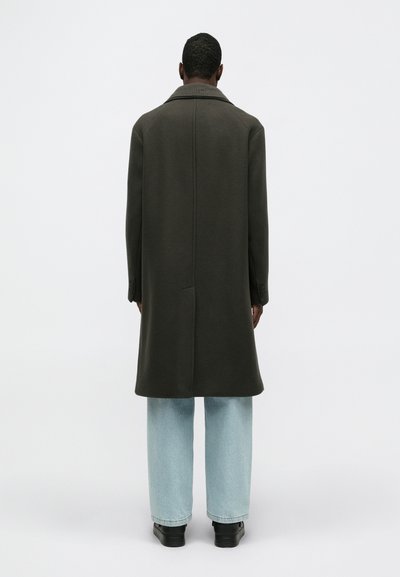 Garšs olīvzaļš mētelis ar ribotu apkakli, taisnas siluets un sānu kabatām, apvienots ar gaiši zilām platām biksēm un melniem apaviem.