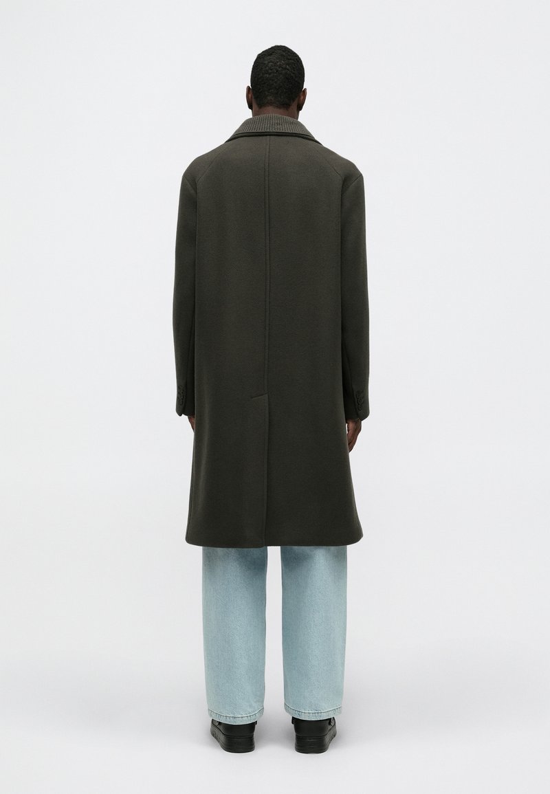 Lang olijfgroen jasje met een ribbelkraag, rechte silhoutte en zijzakken, gecombineerd met lichtblauwe wijde jeans en zwarte schoenen.