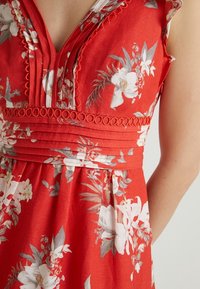 Červené květinové šaty s véčkovým výstřihem, skládacími průramky a kulatými ozdobnými prvky. Materiál je lehký s bílými a šedými květinovými vzory.