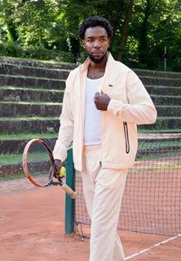 Man in a light tan tracksuit with a zip-up jacket and matching pants, holding a tennis racket and ball on a clay court. Green background.