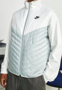 Person som bär en vit och ljusgrå quiltad väst från Nike över en vit långärmad tröja, stående inomhus med händerna i fickorna.