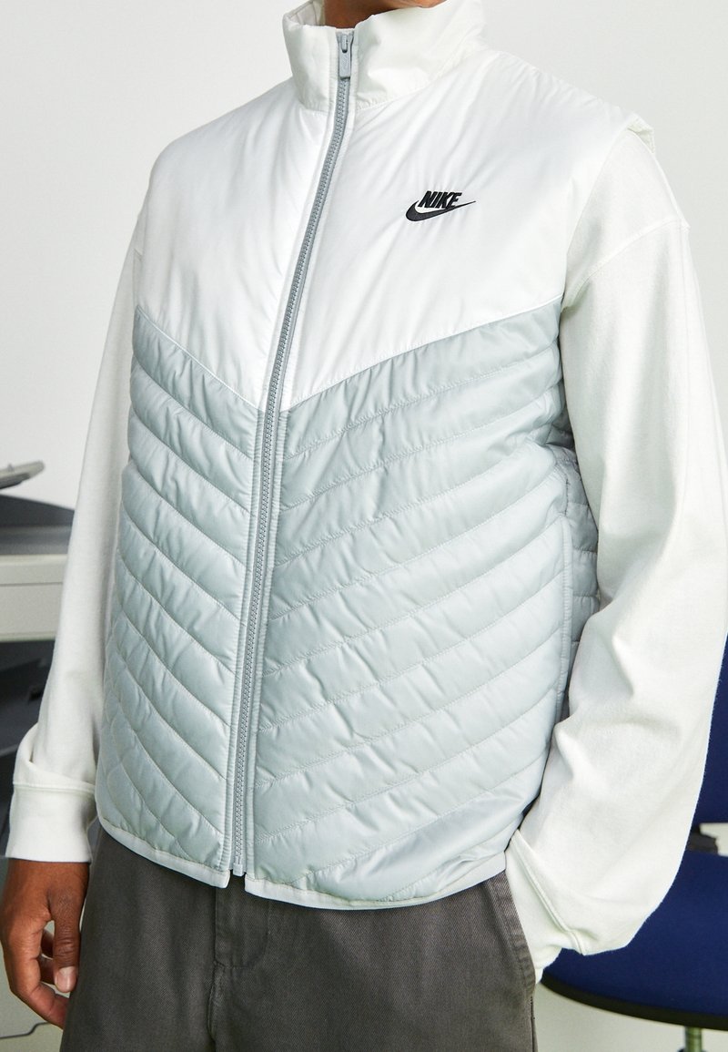 Person som bär en vit och ljusgrå quiltad väst från Nike över en vit långärmad tröja, stående inomhus med händerna i fickorna.