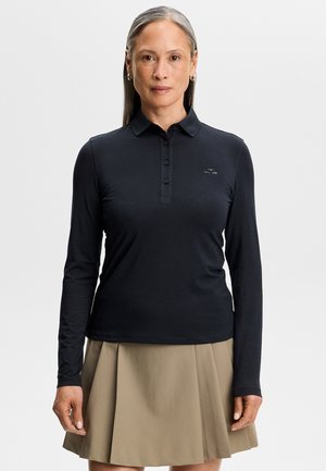 Donna con i capelli lunghi e grigi che indossa una polo nera a maniche lunghe e una gonna beige plissettata, in piedi contro uno sfondo semplice.