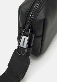 Bolso de cuero negro tipo bandolera con textura suave, que cuenta con un cierre de cremallera y una etiqueta metálica de Calvin Klein sujeta a la correa.