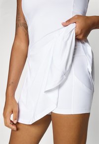 Moteris baltuose sportiniuose šortuose numačius juos, kad parodytų po jais esančius šortus, turinti tatuiruotes ant rankos ir delno.