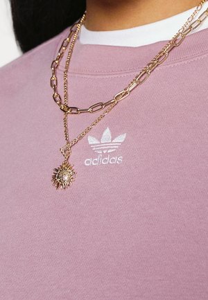 Gros plan d'une personne portant plusieurs colliers en chaîne d'or superposés sur un sweat-shirt rose Adidas avec logo blanc et col de sous-vêtement blanc visible.