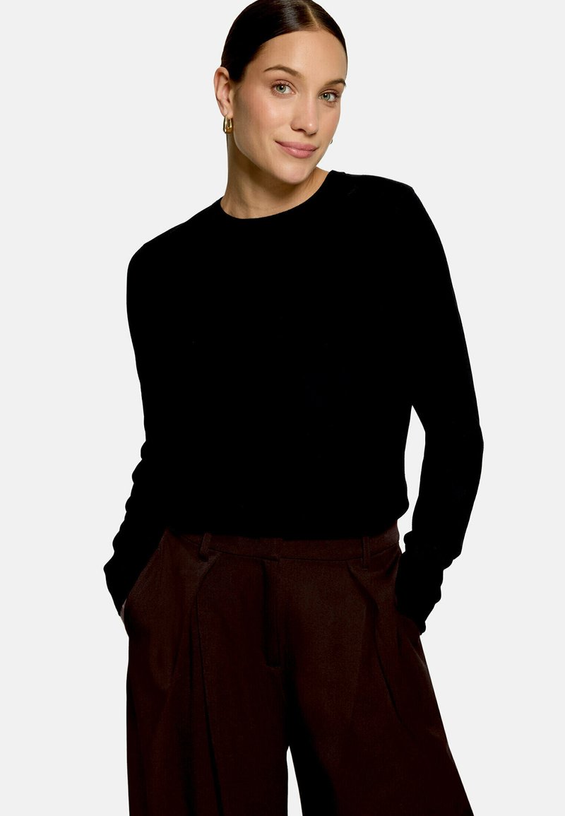 Femme aux cheveux foncés portant un haut noir à manches longues et un pantalon marron foncé, debout les mains dans les poches, souriant légèrement sur un fond uni.