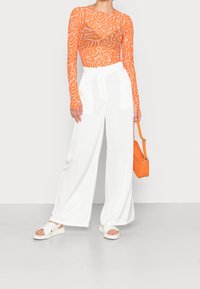 Oranje top met lange mouwen en abstracte spiraalprint, gecombineerd met witte broek met wijde pijpen en witte sandalen met kruisbandjes, met een oranje tas in de hand.