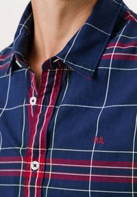 Zbliżenie na osobę w granatowo-czerwonej koszuli w kratę z białymi przeszyciami i małymi czerwonymi haftowanymi inicjałami "PM."