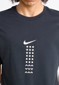 Juodas medvilninis T-palaidinė su centriniu baltu Nike logotipu ir vertikaliu įvairių geometrinių formų išdėstymu balta spalva.