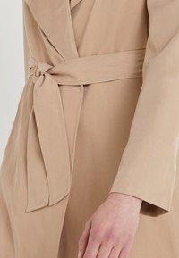 Beige omslagsjacka med knytband, mjuk textur, långa ärmar och subtila sömnadsdetaljer längs kanterna och kragen.