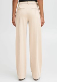 Beige weit geschnittene Hosen mit glatter Textur, ausgestattet mit einer hohen Taille, Gesäßtaschen und scharfen Falten für einen maßgeschneiderten Look.
