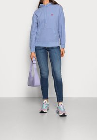 Person med ljuslila hoodie, blå skinny jeans, iriserande sneakers, som håller en lavendelfärgad handväska mot en enfärgad bakgrund.