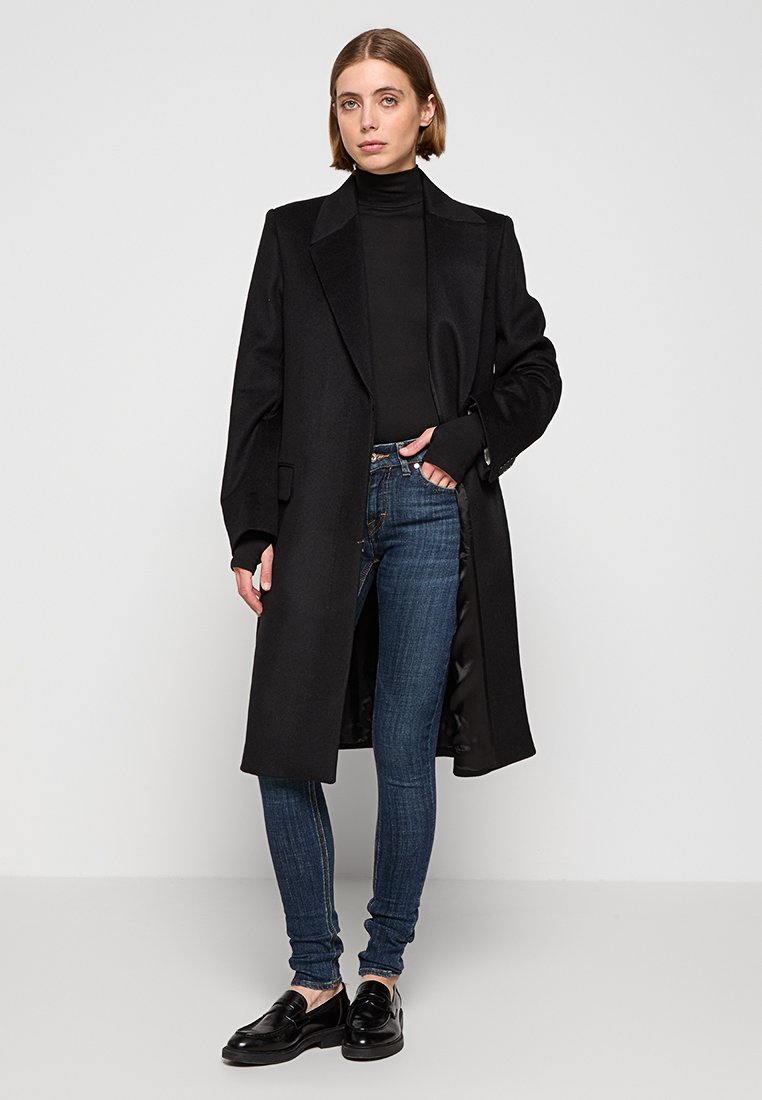 Manteau long en laine noire avec des revers en pointe, accompagné d'un pull à col roulé noir et d'un jean skinny bleu foncé. Assorti à des mocassins noirs.