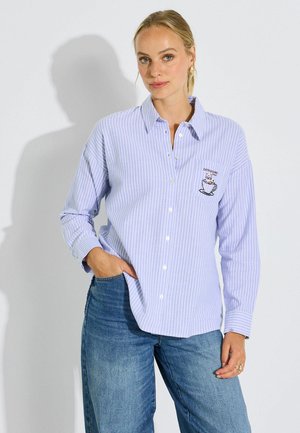 Mujer rubia con camisa de botones a rayas azul claro con diseño bordado de gato en taza de café y jeans azules, mano en el bolsillo.