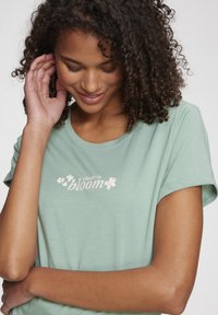 Mentolovo zelené tričko s krátkymi rukávmi, ktoré obsahuje nápis "vyber sa rozkvetnúť" a kvetinové motívy v svetloružovej farbe. Mäkká štruktúra látky.