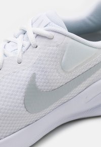 Chaussure de sport blanche avec une tige en mesh texturé, des accents ton sur ton et un swoosh Nike argenté réfléchissant. Les lacets sont intégrés avec un léger rembourrage.