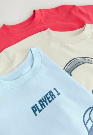 Tre crew neck sweatshirts i rød, creme og lyseblå. Hver har ribbede manchetter, med "PLAYER 1" trykt på den blå.