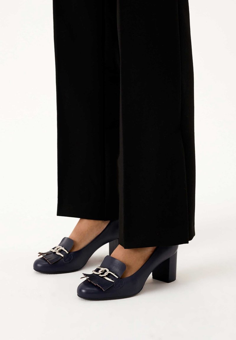 Femme portant un pantalon large noir et des mocassins à talons carrés bleu marine avec des détails en chaîne argentée et des pompons.