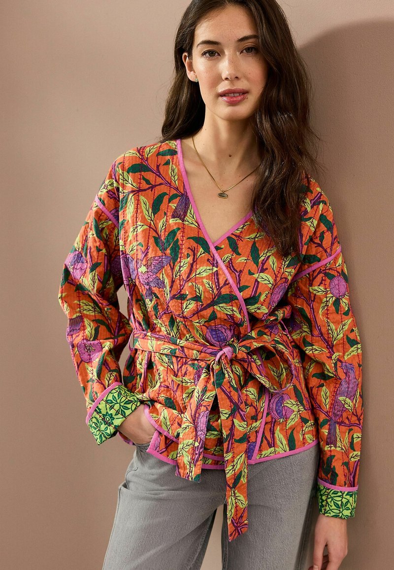 Top envolvente colorido con un fondo naranja y patrones florales en verde y púrpura, decorado con un ribete rosa, combinado con pantalones grises.