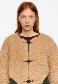 Joven con cabello rojo y pecas, vestida con una chaqueta beige de felpa con tres botones de palanca negros, posando frente a un fondo blanco liso.