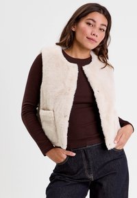 Femme portant un gilet en fausse fourrure beige sur une chemise à manches longues marron foncé et un jean en denim noir, les mains dans les poches, posant contre un fond blanc.