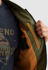 Main tenant une veste verte et orange ouverte montrant une poche intérieure zippée et un t-shirt bleu marine avec un motif graphique de texte et une carte.