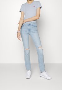 Maglietta a righe blu con piccolo logo rosso, abbinata a jeans strappati azzurri. Indossata con sneakers bianche e un elastico viola al polso.