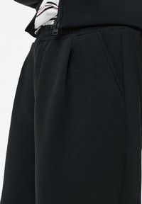 Pantalon noir taille haute avec plis et poches latérales, porté avec une veste noire zippée et une chemise rayée rentrée.