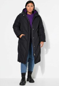 Svart lång pufferjacka med huva, diamantquiltat mönster, frontdragkedja och sidofickor. Bärs över en lila tröja och jeans.