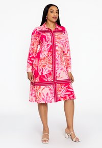 Knopfkleid mit einem lebhaften rosa- und orangefarbenen Blumenmuster, langen Ärmeln, Kragen und knielangem Saum. Kombiniert mit weißen Sandalen.