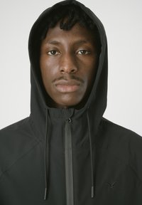 Schwarze Kapuzenjacke mit Reißverschluss, glatte Textur, ausgestattet mit verstellbaren Zugbändern und dezentem Logo-Detail auf der Brust.