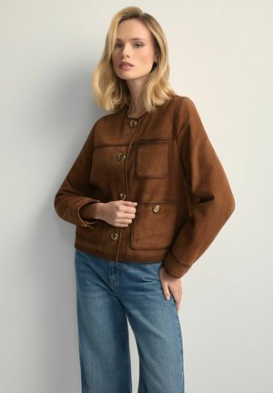 Kobieta w brązowej zamszowej kurtce zapinanej na guziki z przednimi kieszeniami i niebieskich dżinsach, stojąca na tle jednolitego jasnoszarego tła.