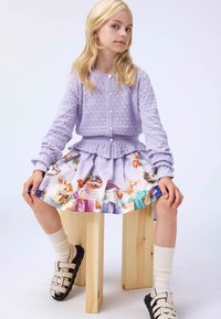 Lila gestrickte Strickjacke mit Rundhalsausschnitt und Perlenknöpfen, kombiniert mit einem plissierten Rock mit bunten Tiermotiven und cremefarbenen Socken.