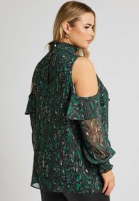 Grön mönstrad blus med cold-shoulder design, volangärmar och smockad halsringning. Genomskinligt tyg med en lätt lyster.
