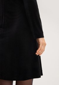 Schwarzes Samtkleid mit einer weichen Textur, langen Ärmeln und einem ausgestellten Saum. Die Hand ist entspannt neben dem Kleid. Minimale sichtbare Details.