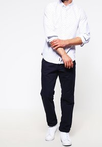 Weißes Langarm-Hemd mit blauen Polka-Dots, aufgerollte Bündchen, kombiniert mit marineblauen Hosen und weißen Sneakern. Glatter Stoff, taillierte Passform.