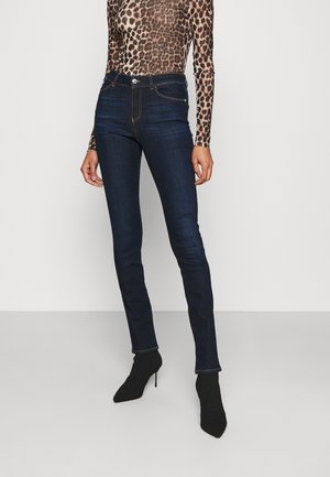 Dark blue skinny jeans with a high waist, featuring orange stitching and a front pocket. Worn with a fitted leopard print top and black heels.