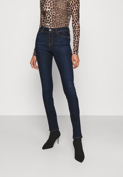 Sötétkék, slim fit farmernadrág magas derekú fazonnal, narancssárga varrással és elülső zsebbel. Fekete magassarkú cipővel és testhezálló leopárdmintás felsővel viselve.