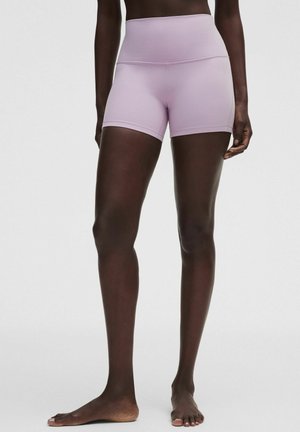 Modelka nosi fioletowe sportowe szorty z wysokim stanem, stoi na boso na jednolitym białym tle.