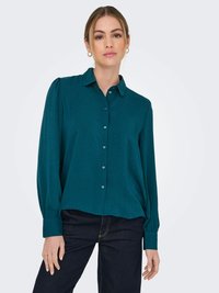 Camicia azzurra con bottoni e maniche lunghe e voluminose, con colletto e tessuto strutturato. Indossata con jeans scuri, che mostrano una vestibilità rilassata.