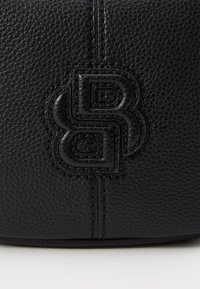 Primo piano di una borsa a tracolla in pelle nera con superficie texturizzata, caratterizzata da un logo in rilievo. Le cuciture sono visibili, aggiungendo dettaglio al design.