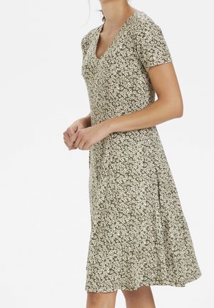 Femme portant une robe courte à manches courtes, encolure en V, longueur aux genoux, avec un motif floral vert et blanc, debout les mains jointes.
