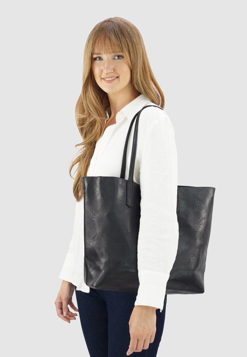 Zwarte leren tote bag met een gestructureerde oppervlakte, rechthoekige vorm en lange hengsels. Wordt gedragen over de schouder door een model in een wit shirt.