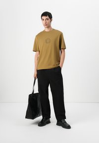 Khaki short-sleeve T-shirt with a smiley face print, paired with black wide-leg trousers and a glossy black tote bag.