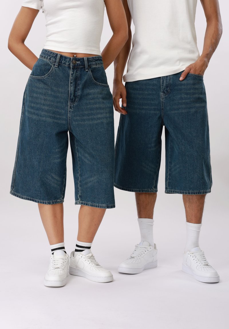 Δύο άτομα που φορούν μακριά τζιν σορτς, λευκά αθλητικά παπούτσια και λευκά τοπ, στέκονται δίπλα δίπλα μπροστά σε ένα απλό φόντο.