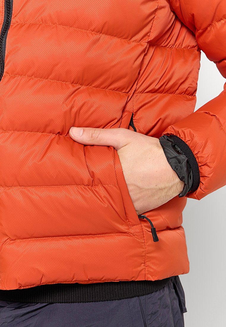 Chaqueta naranja acolchada con diseño de acolchado, bolsillo con cremallera y puños elásticos. La tela parece ligera con una textura suave y brillante.