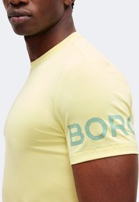 Κοντινή λήψη ενός ατόμου που φοράει ένα ανοιχτό κίτρινο κοντομάνικο πουκάμισο με μερικό μπλε κείμενο "BORG" στο αριστερό μανίκι.